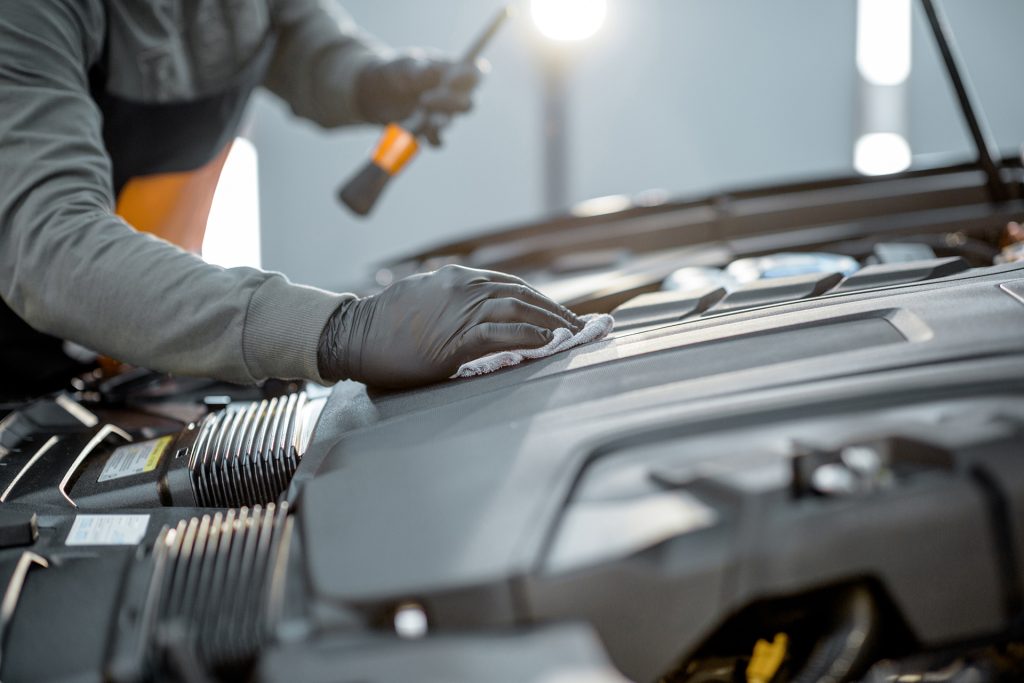 A mechanic wearing gloves inspects a car engine with a flashlight.