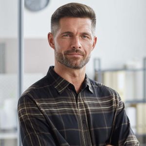 Confident man with a beard in a plaid shirt standing indoors.