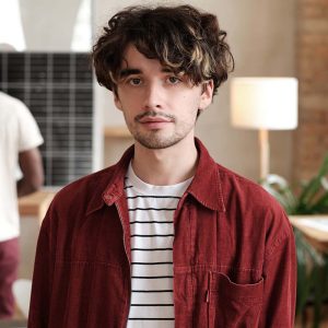 A young man with curly hair wearing a striped shirt and red jacket.